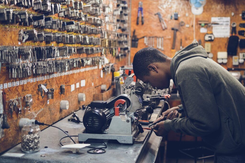 Person using key-cutting machine in a locksmith shop with rows of keys on the wall.