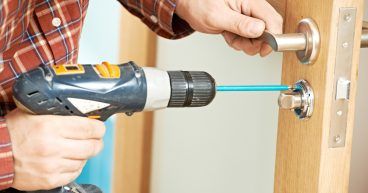 Person using a power drill to install a door handle on a wooden door.
