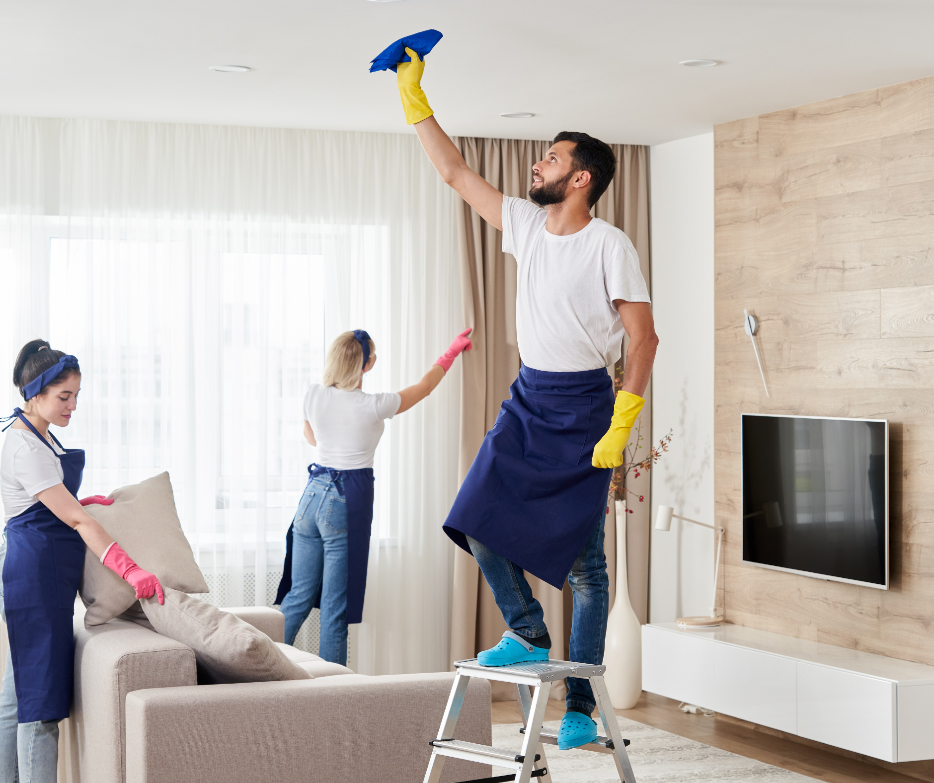Three people cleaning a living room. One on a stepladder cleans the ceiling, two dust furniture.