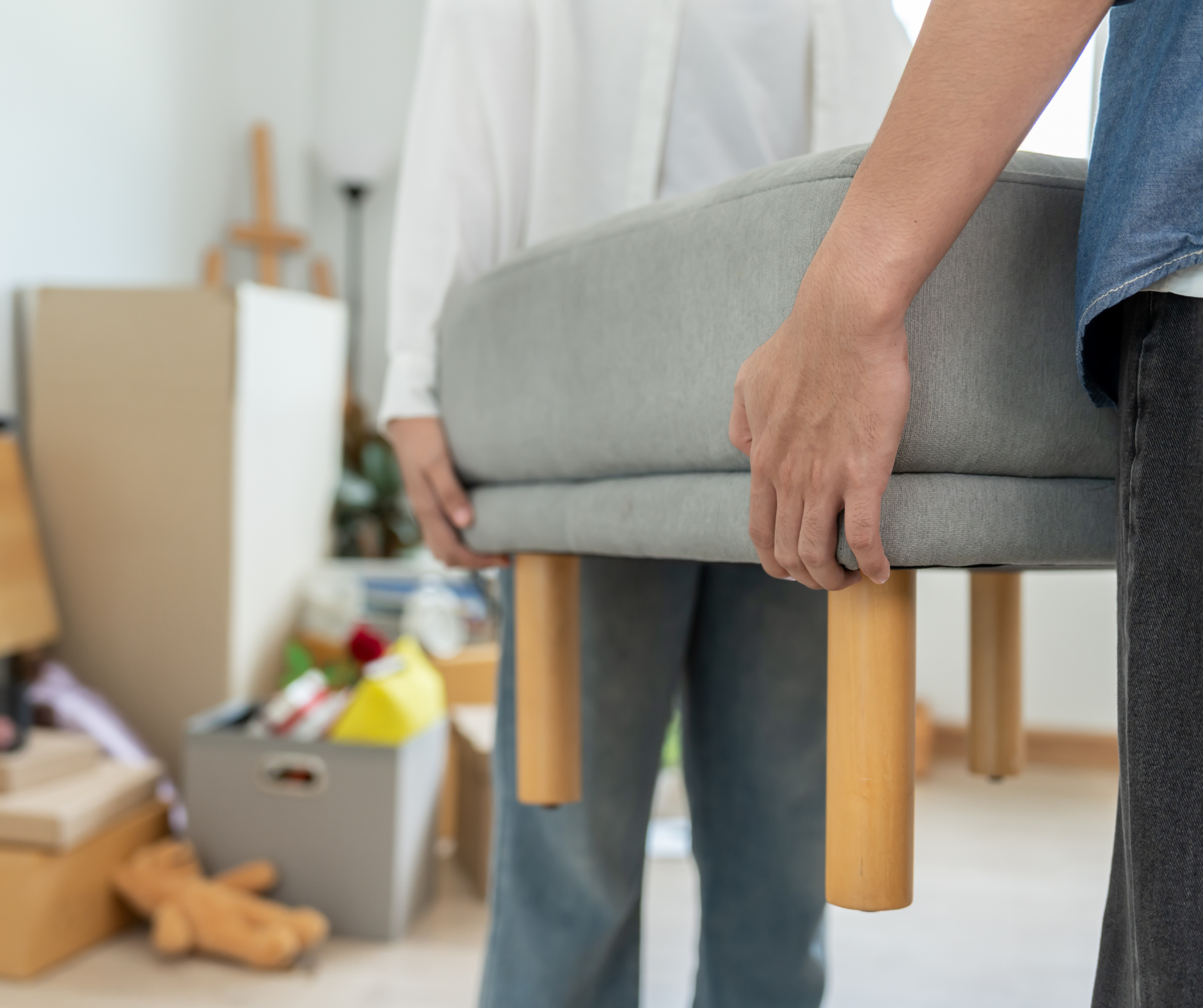 Two people carrying a gray upholstered bench with wooden legs in a room with packed boxes.