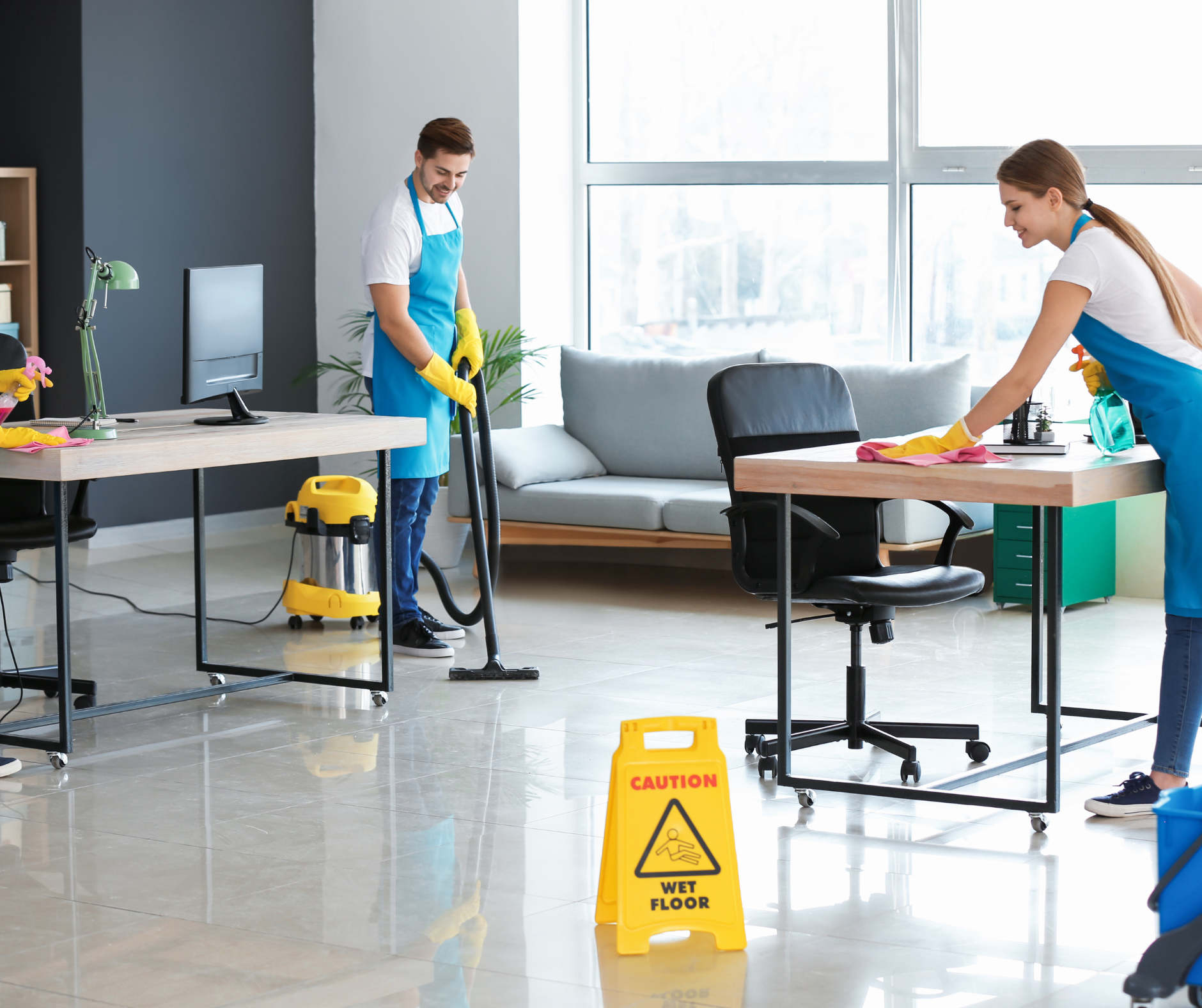 Two people cleaning an office: vacuuming floor and dusting desk. Yellow caution sign on the floor.