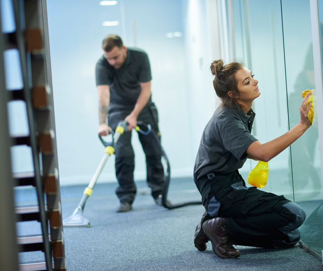 Two people cleaning an office: one vacuuming the carpet, the other wiping a glass wall.