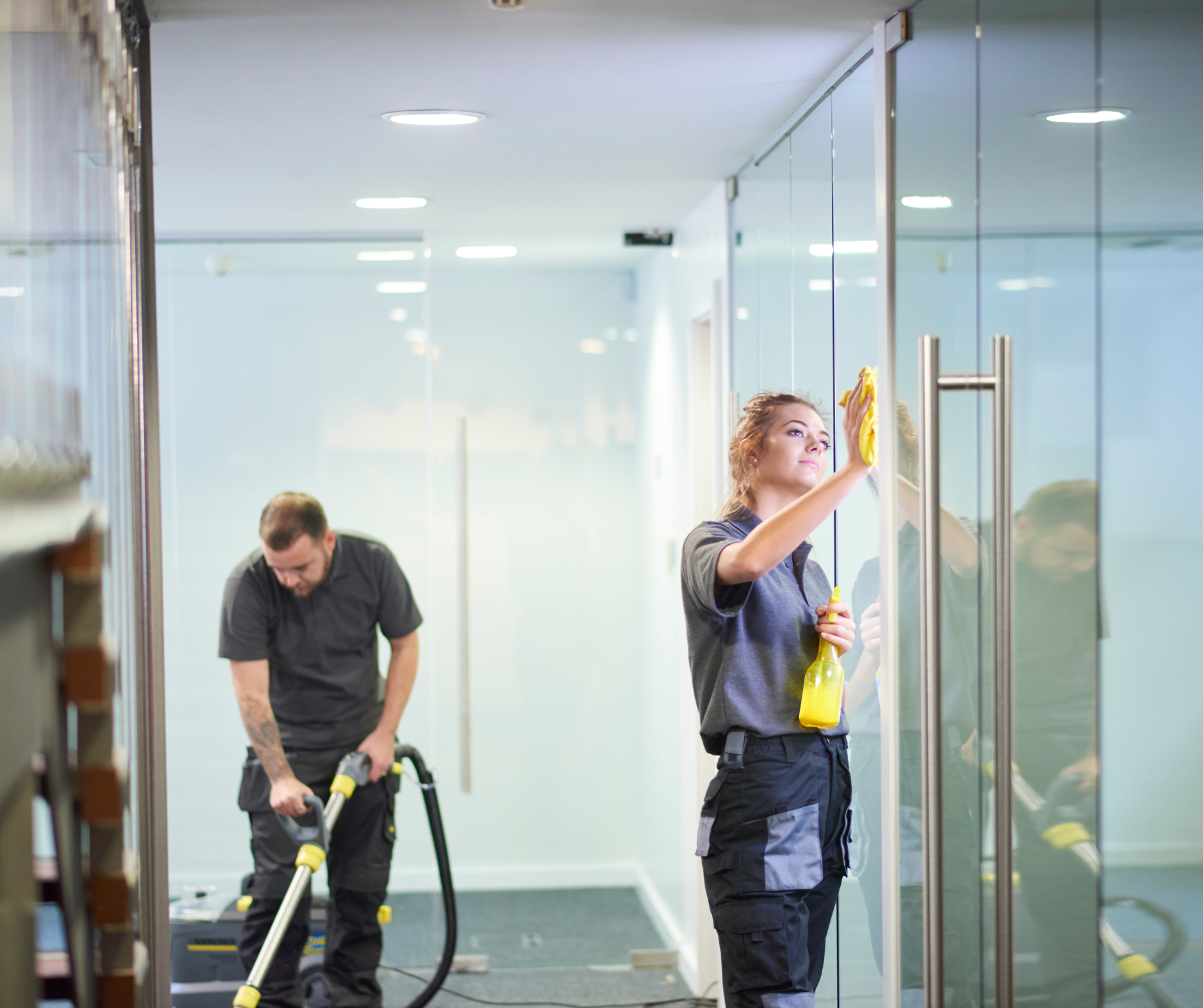 Two people cleaning an office: one vacuuming the floor, the other wiping a glass door.