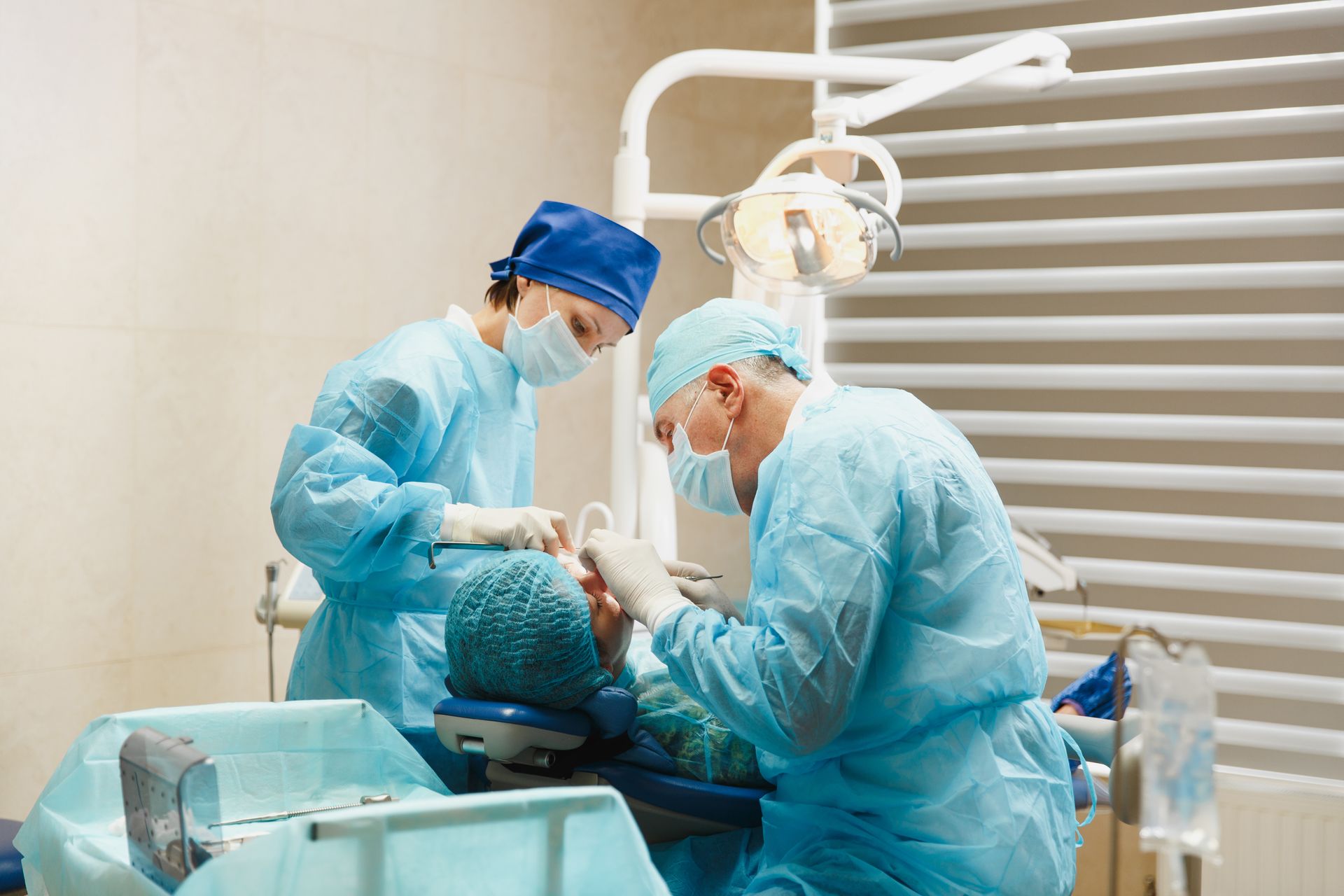 Photograph of two medical professionals wearing blue surgical scrubs, masks, and caps performing a dental procedure. Photograph of two medical professionals wearing blue surgical scrubs, masks, and caps performing a dental procedure.