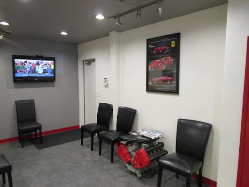 Waiting room with chairs, TV, and a car engine. Poster of a Ferrari on the wall. Grey and white walls.