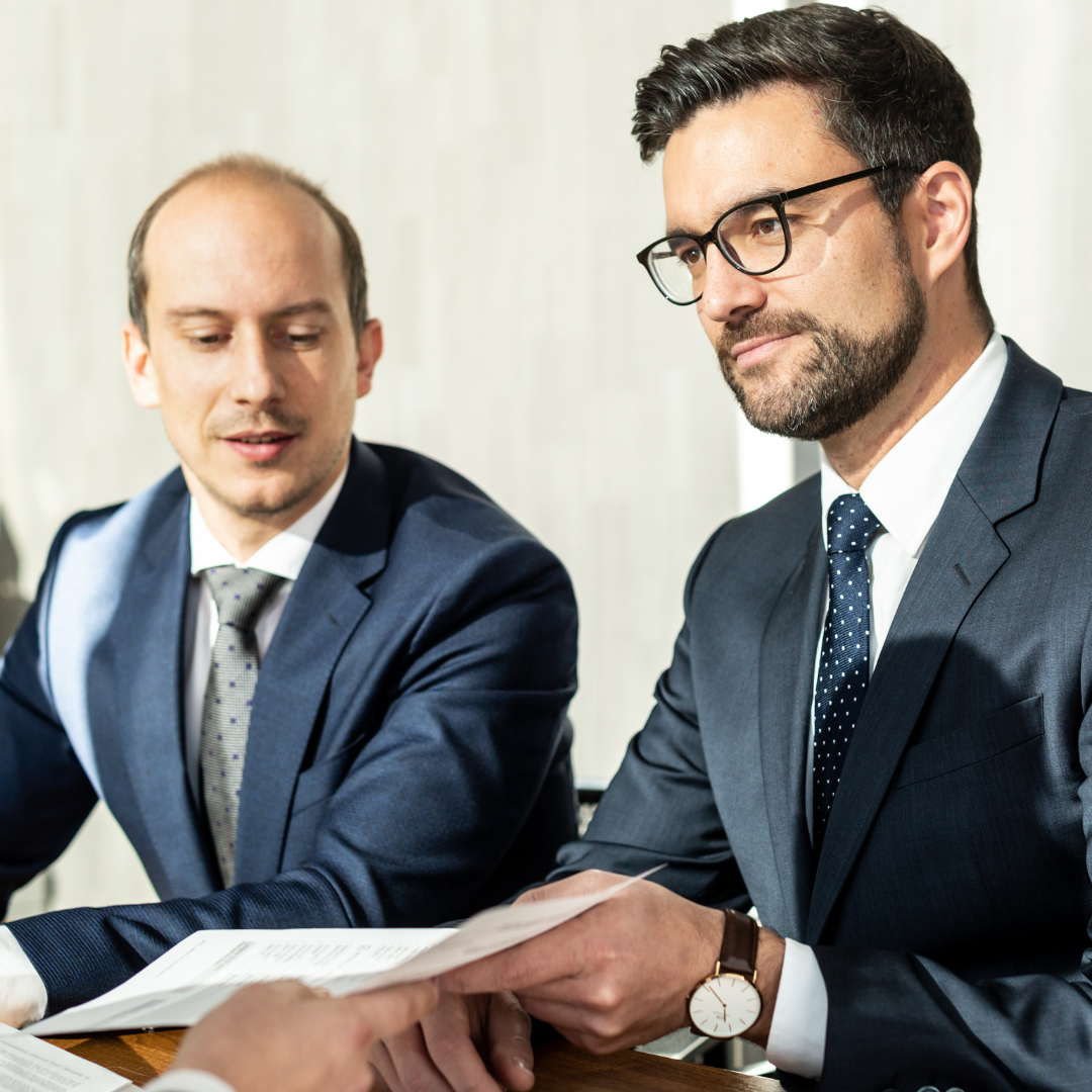 Two men in suits and ties are sitting at a table looking at papers