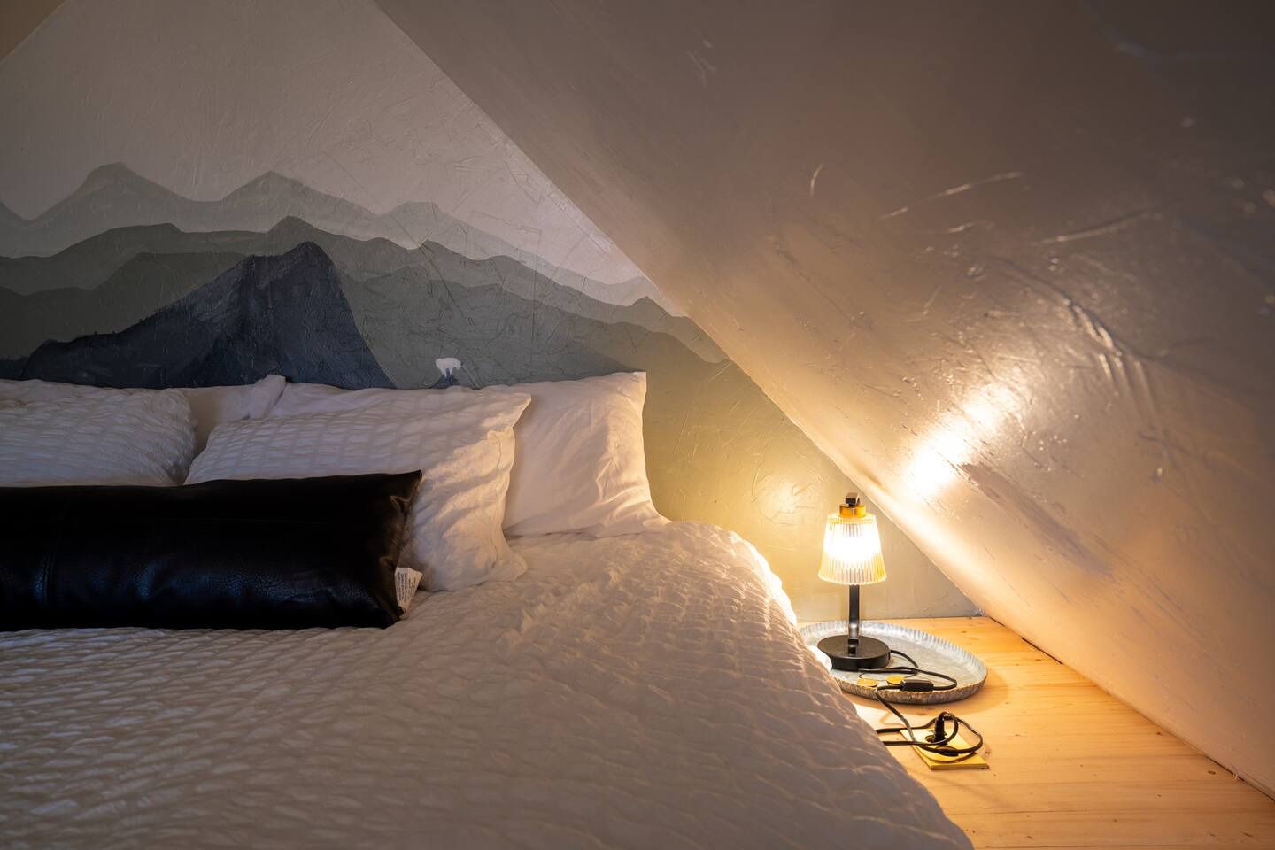 There is a bed in the attic with a painting of mountains on the wall.