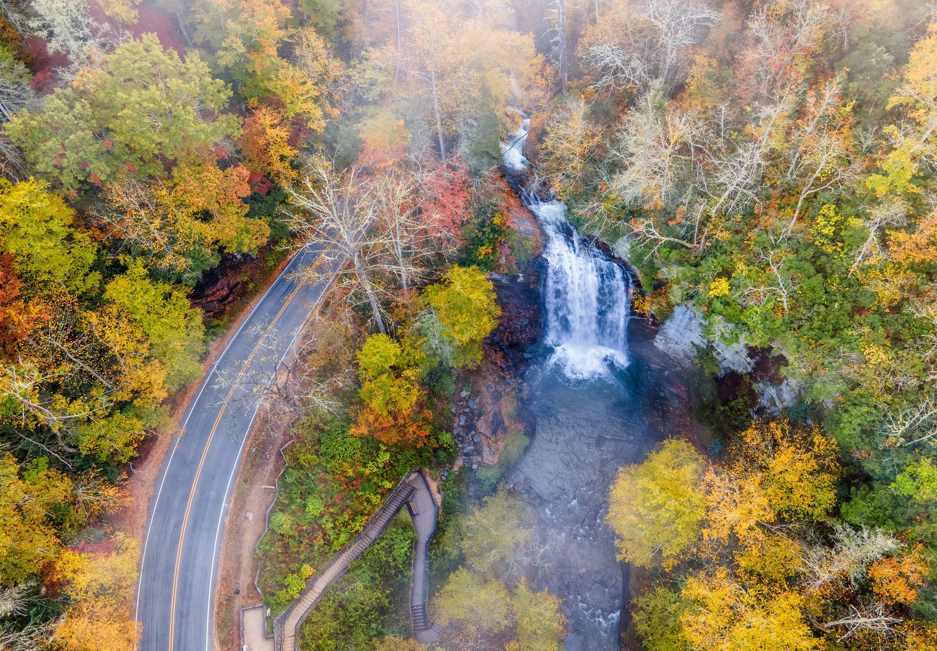 Aerial view of a waterfall and winding road surrounded by colorful autumn trees.