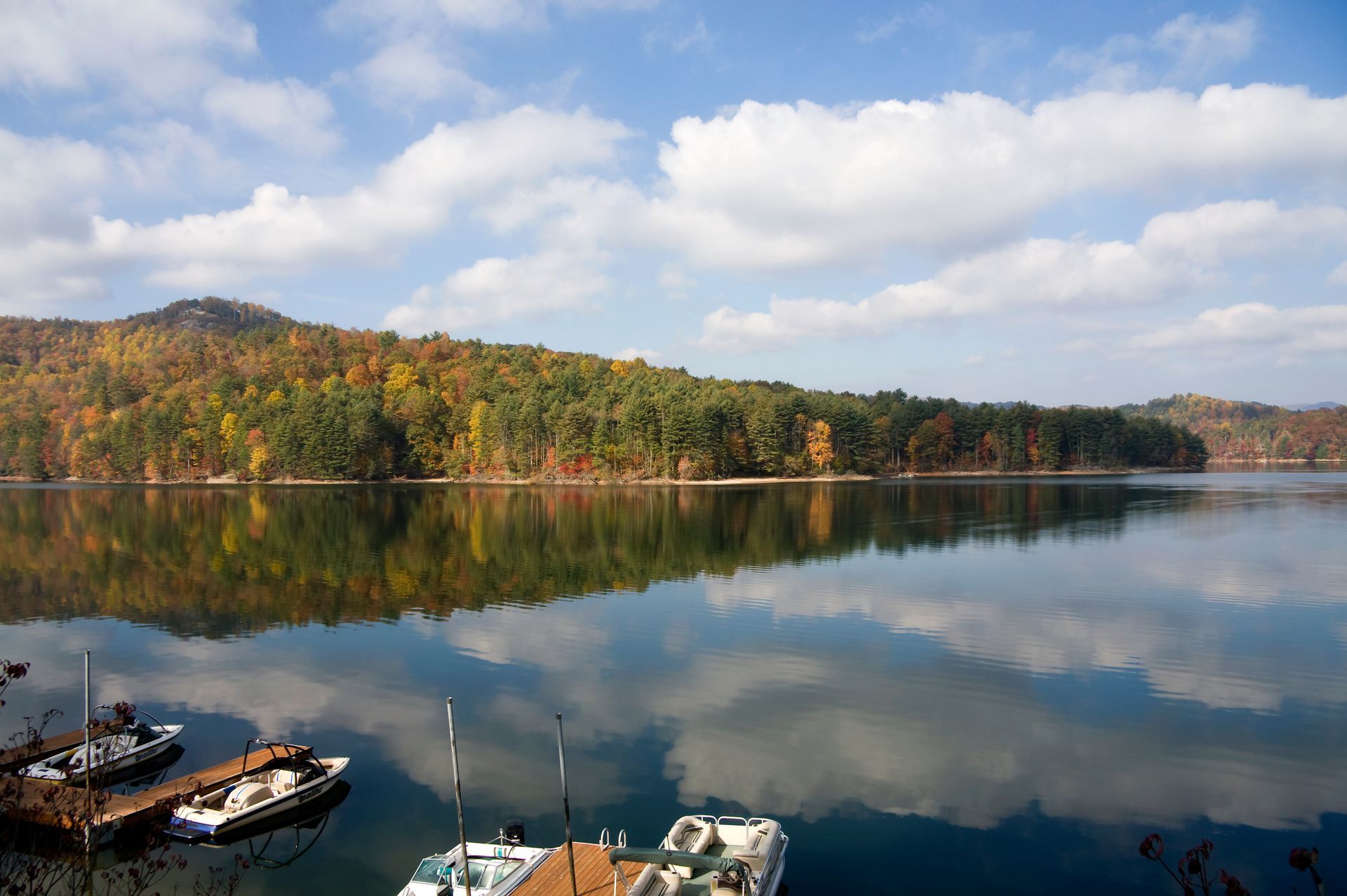 Still lake reflects trees in autumn colors and blue sky; docks with boats.