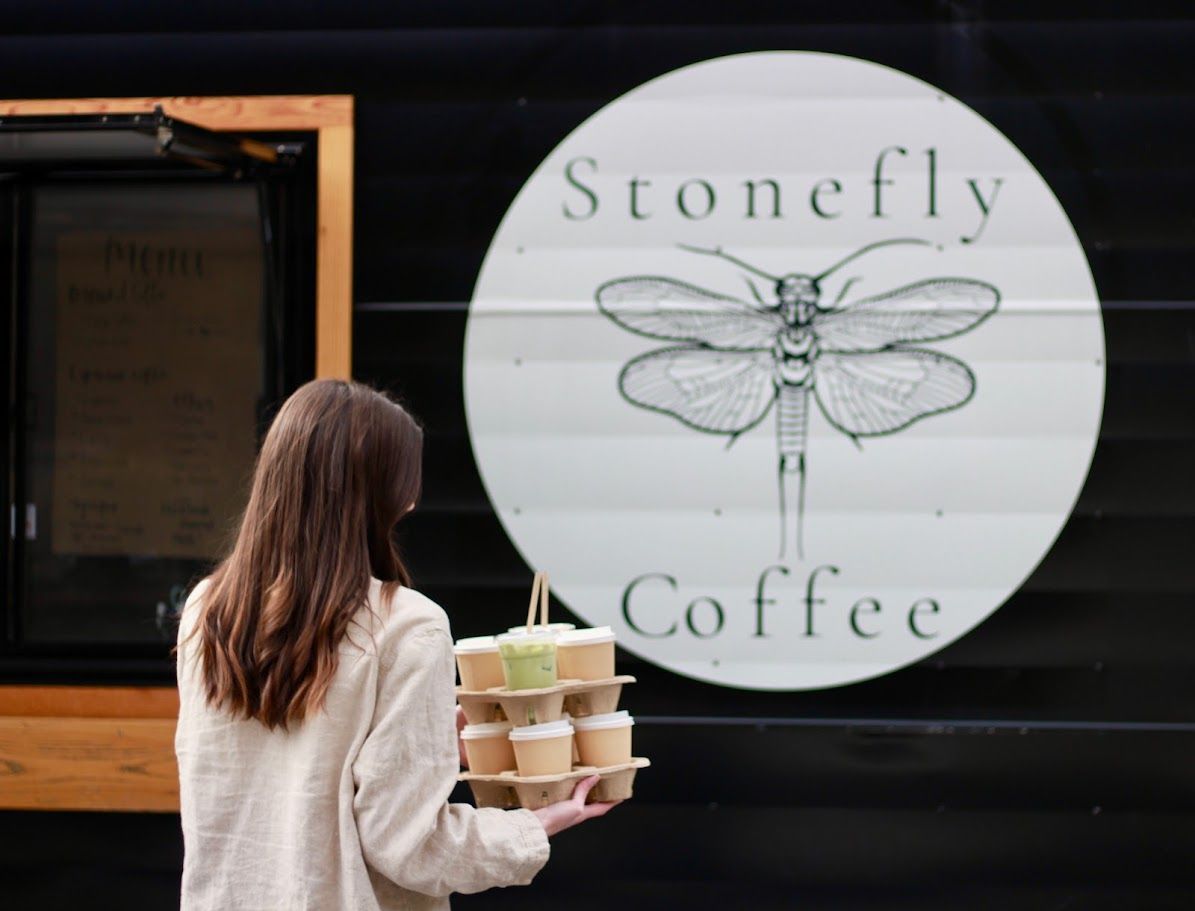 A customer receiving a coffee from a food truck, with a wooden counter and menu visible.