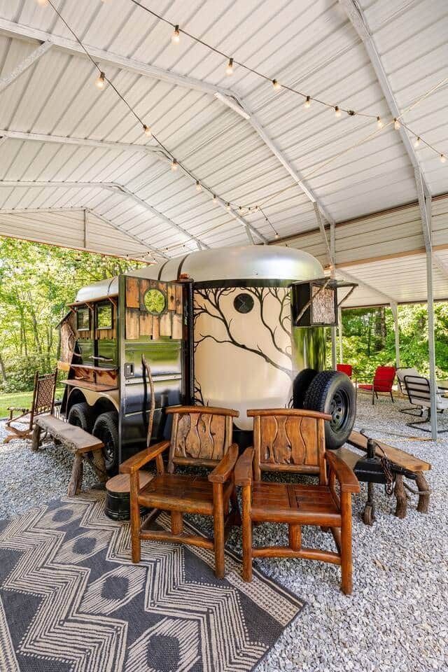 A horse trailer is parked under a covered area with chairs and a rug.