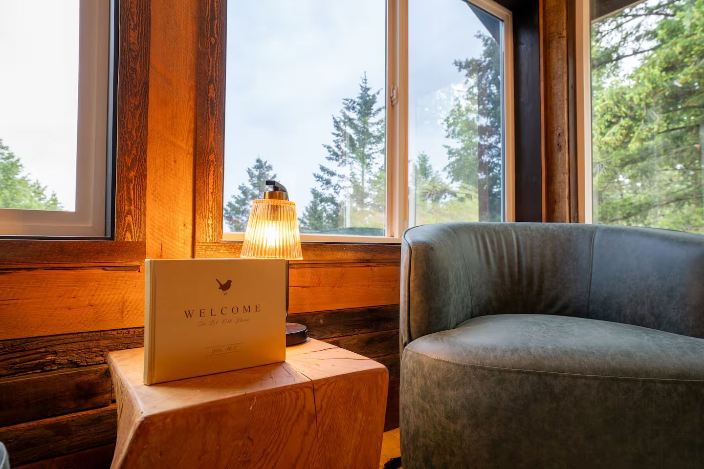 A book is sitting on a wooden table next to a chair in front of a window.