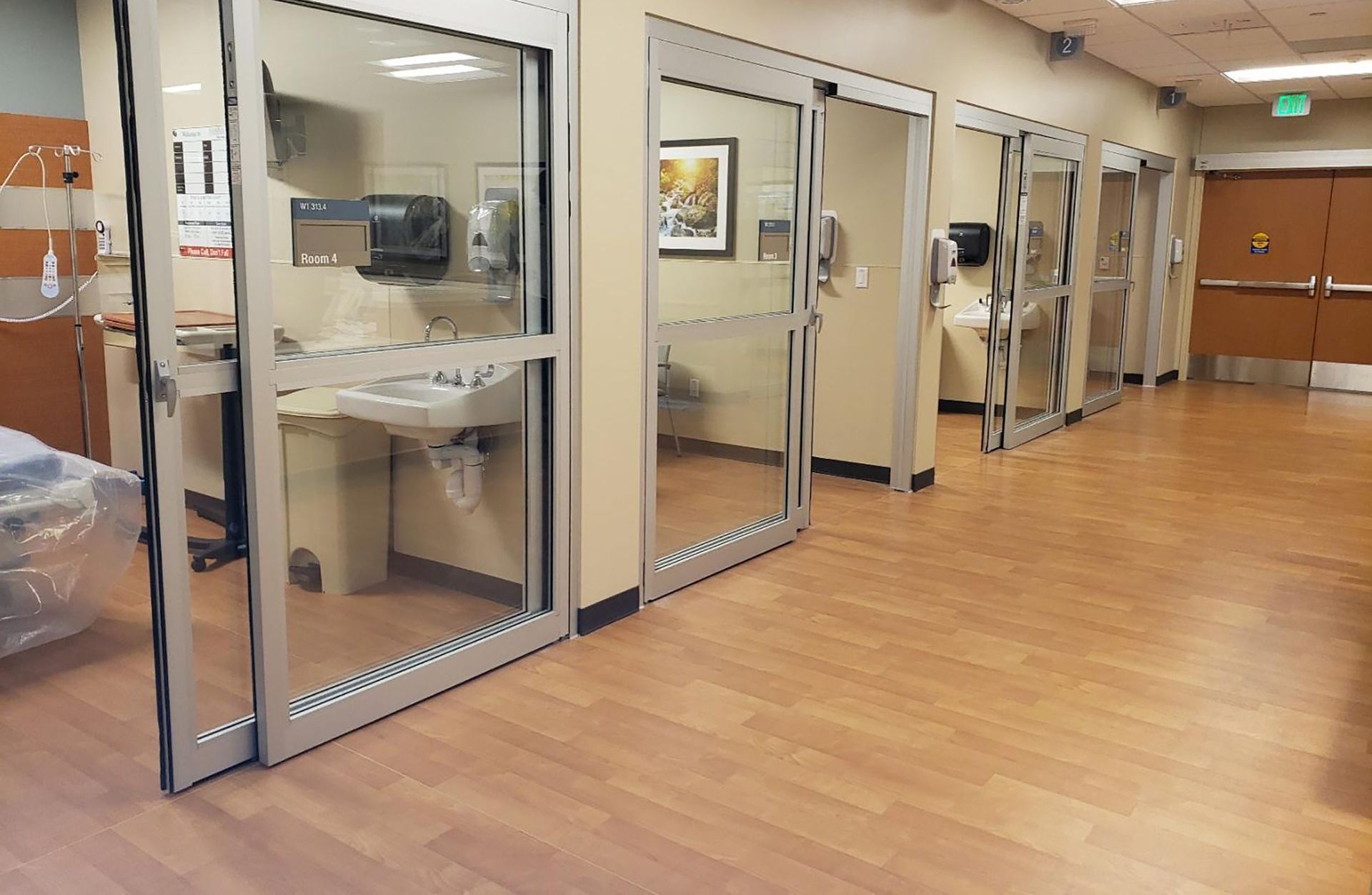 Couloir d'hôpital avec portes vitrées et salles d'examen, doté d'un parquet et de murs beiges.