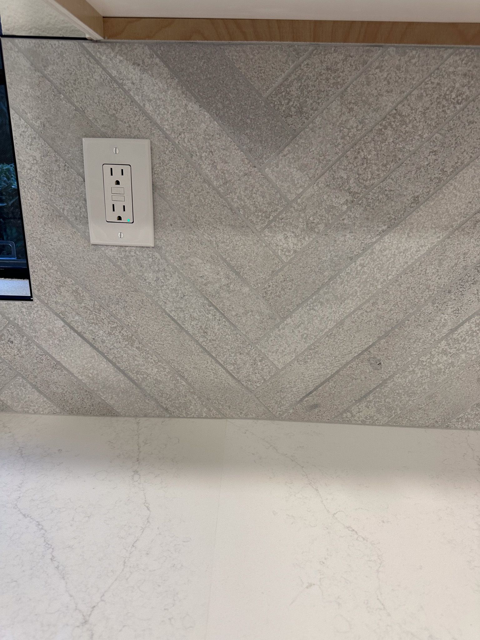 A close up of a gray tile wall with a white electrical outlet.