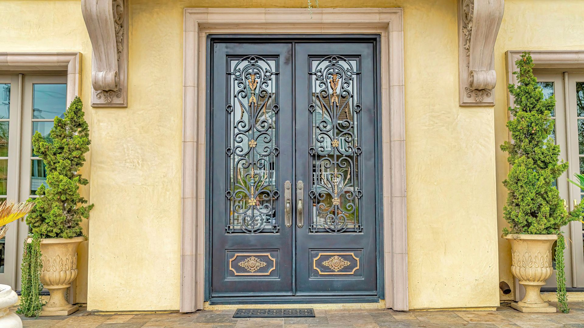Double black ornate front doors with glass panes, flanked by potted topiaries and windows on a beige stucco facade.