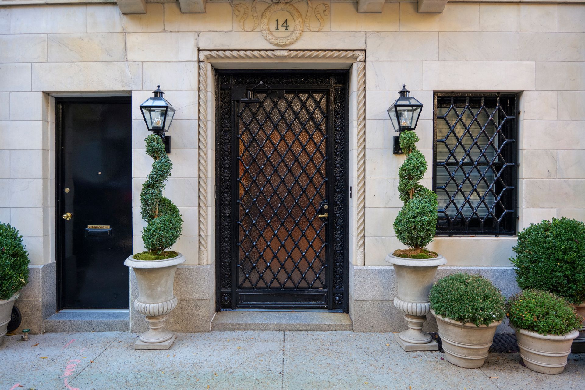 Exterior view of building with ornate black door and security grid, flanked by topiary and sconces.