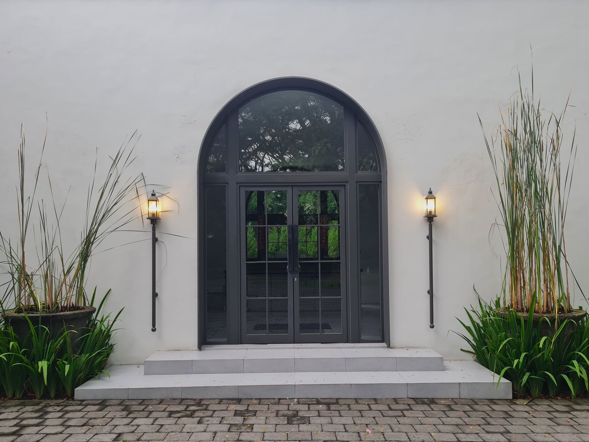 A dark arched doorway with glass panels, flanked by lights and potted plants, against a white wall.