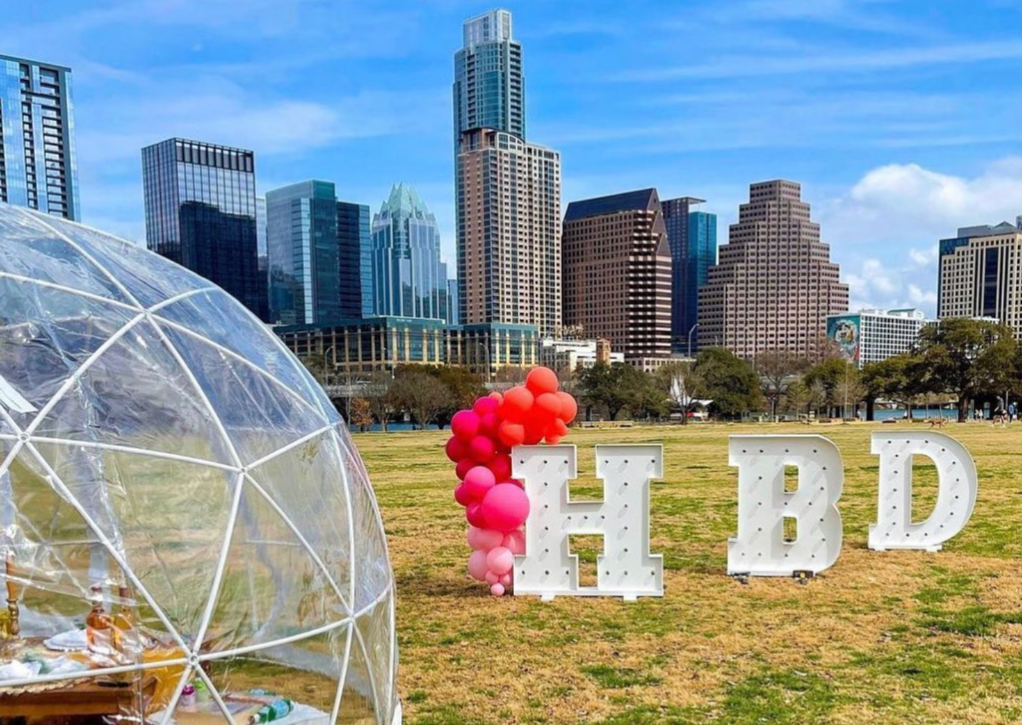 A field with balloons and letters that say hbd in front of a city skyline.