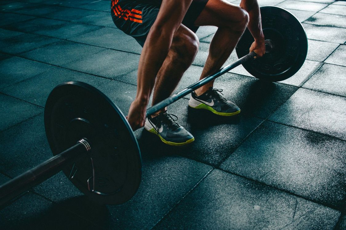 Una persona solleva un bilanciere sul pavimento di una palestra, indossando scarpe fluorescenti in uno spazio per l'allenamento scarsamente illuminato.