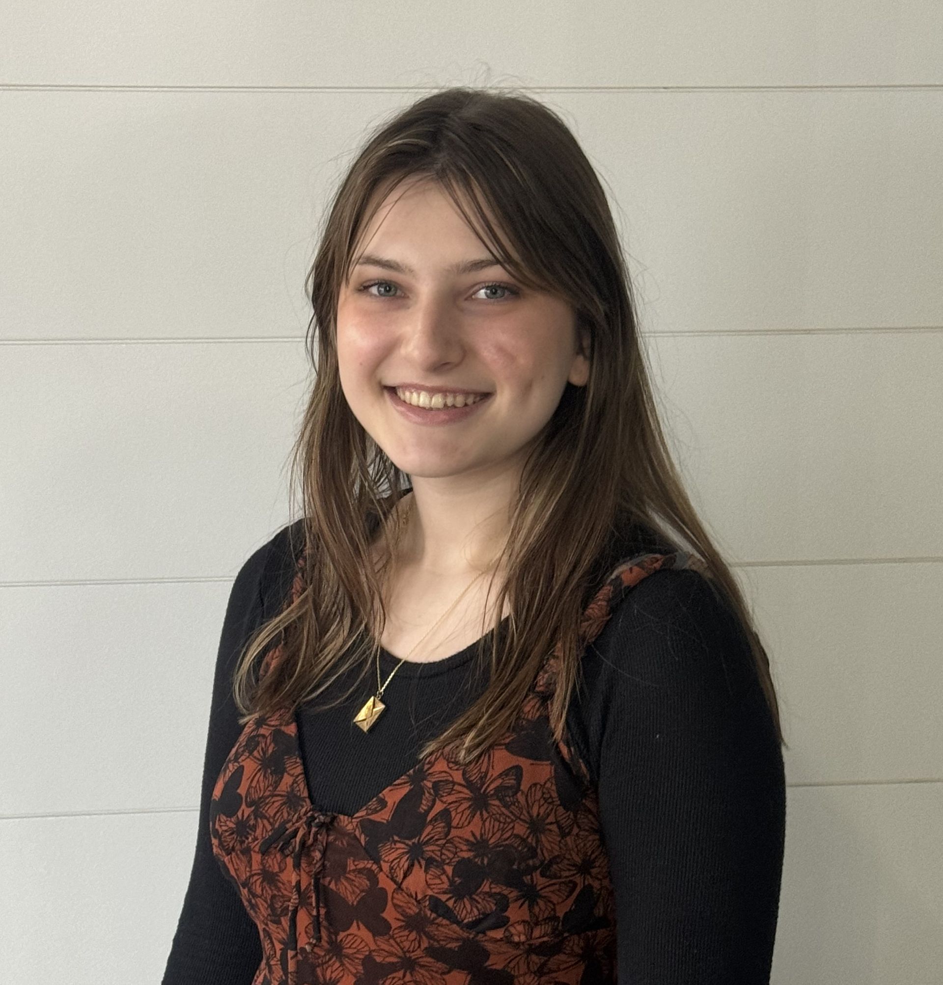 Woman smiling, wearing a black top and patterned dress, in front of a white paneled wall.