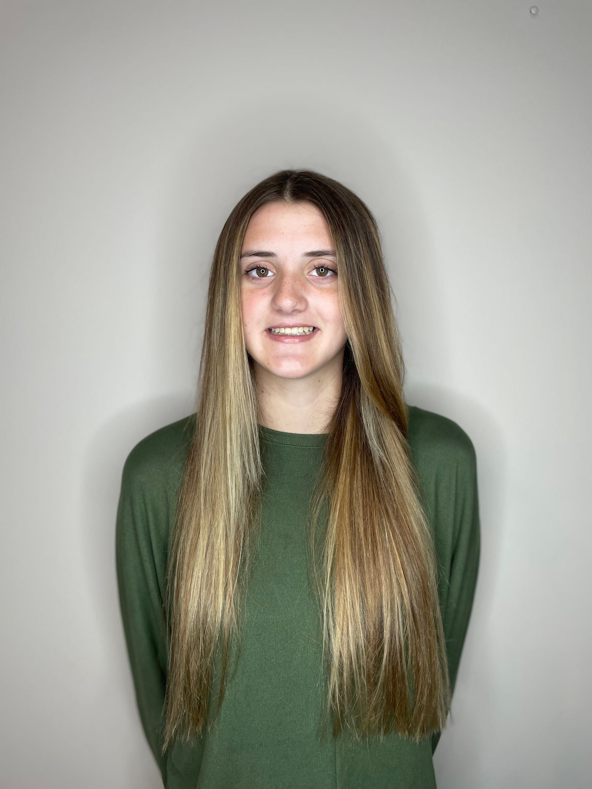 Woman with long, blonde and brown hair smiling, wearing a green long-sleeved shirt against a gray background.