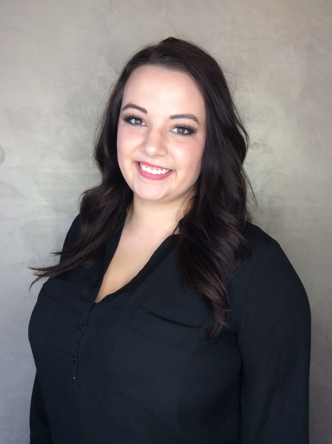 Woman with dark wavy hair smiles, wearing a black blouse against a gray backdrop.