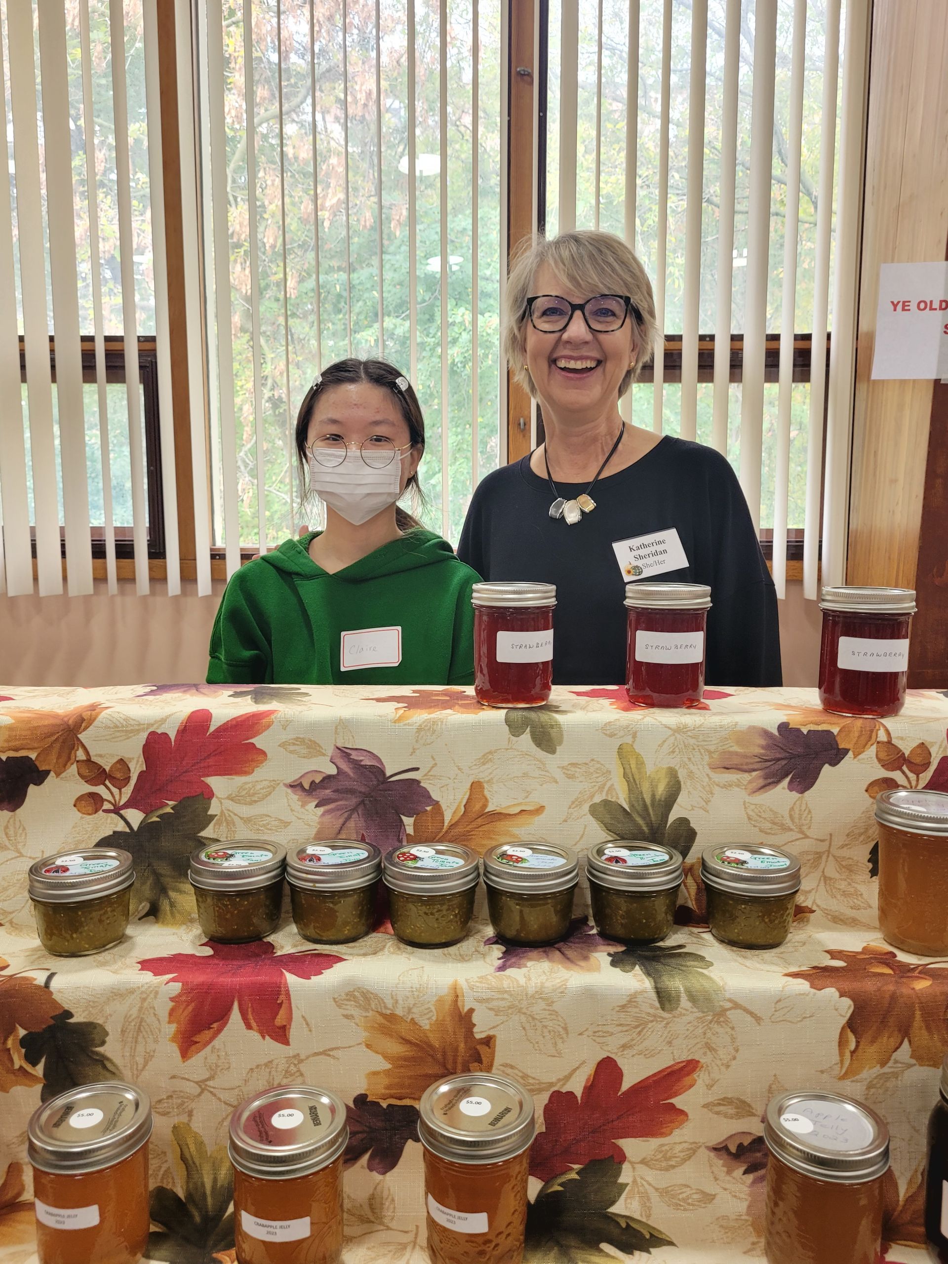 a woman and a girl are standing in front of a table filled with jars of jam .