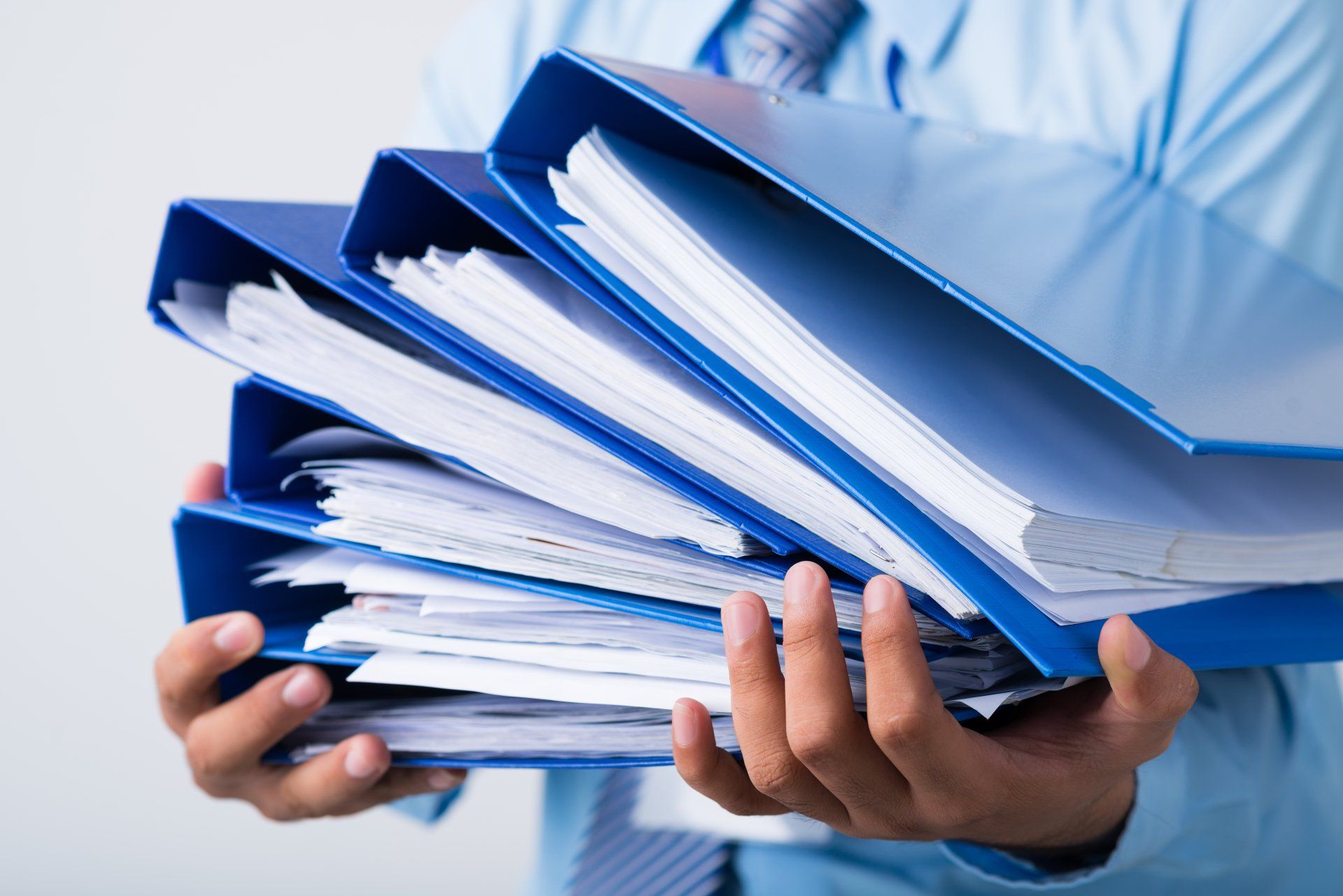 a man in a blue shirt and tie is holding a stack of blue binders filled with papers .