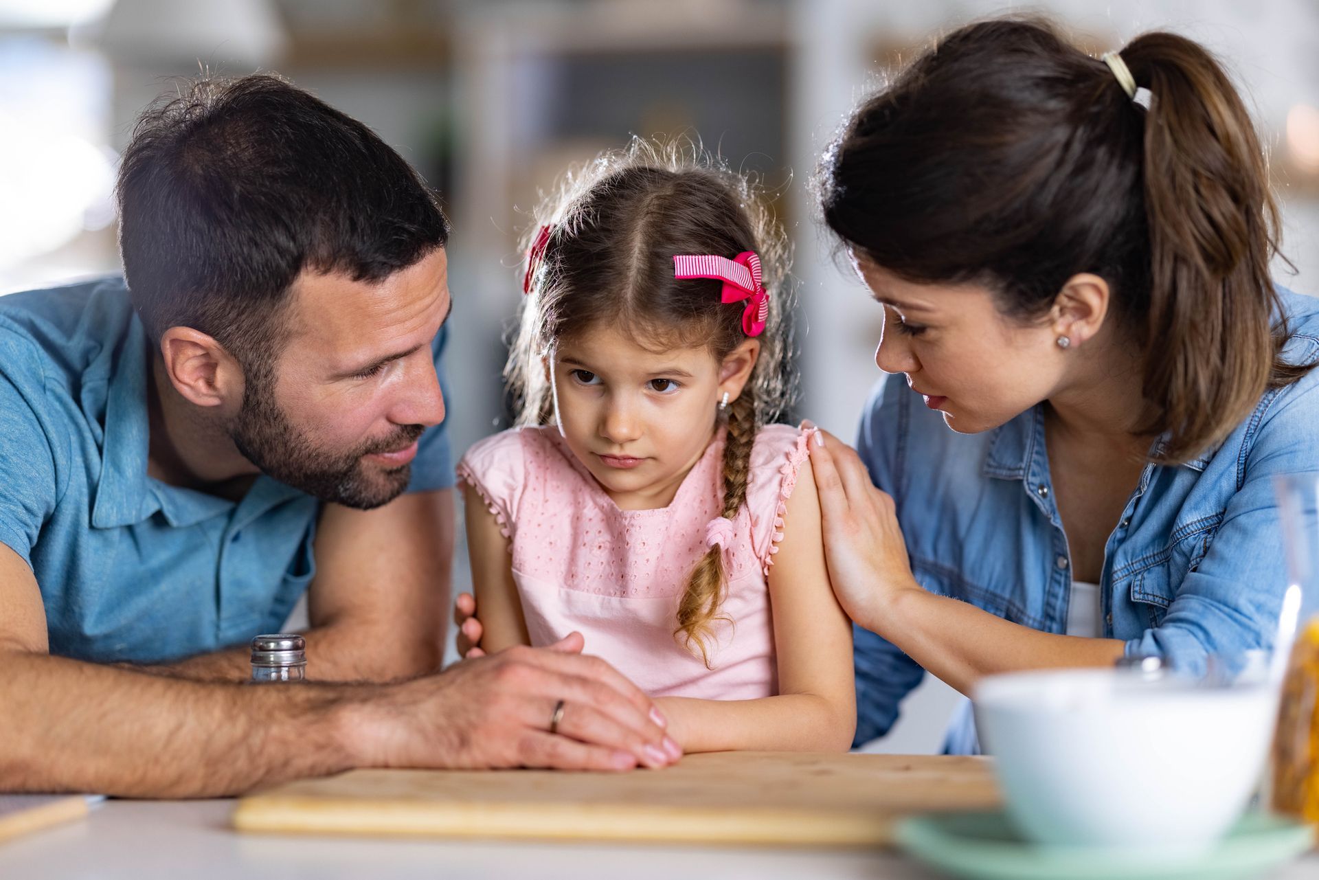 A man and woman are comforting a little girl who is sitting at a table.