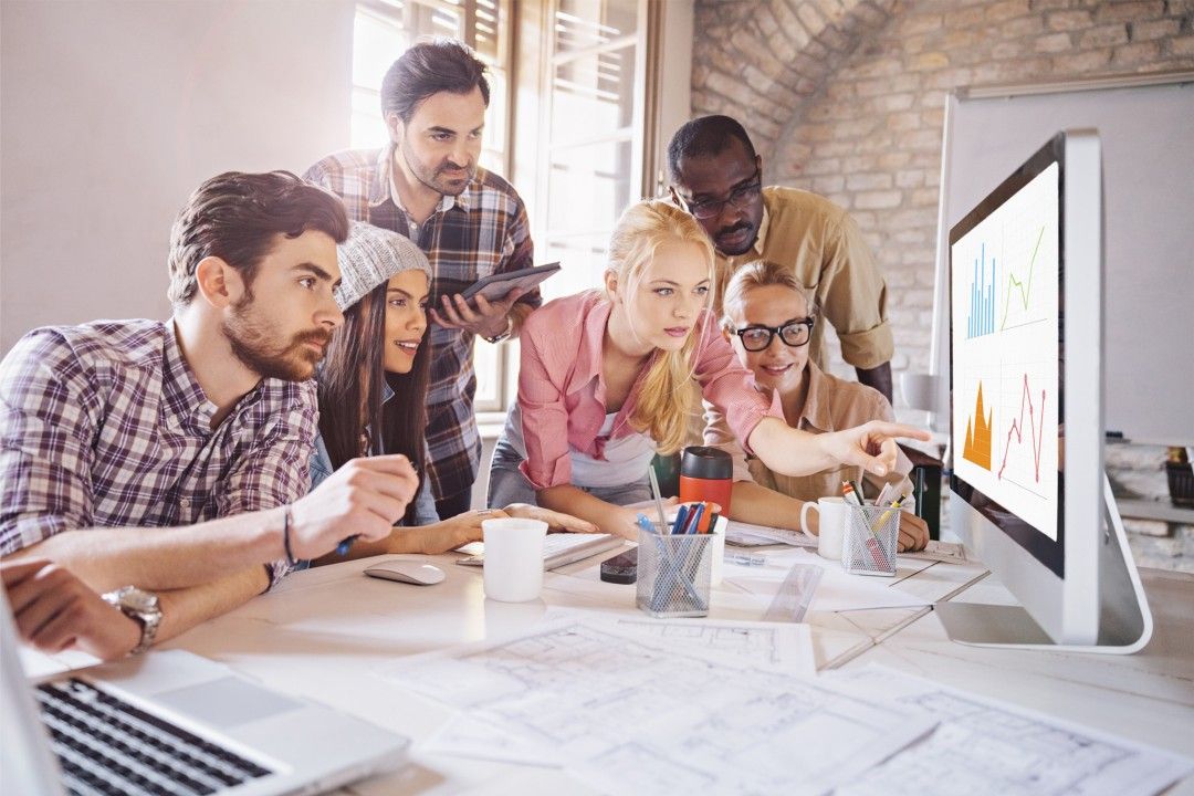 Team of six colleagues looking intently at a computer screen, discussing charts and graphs in an office.