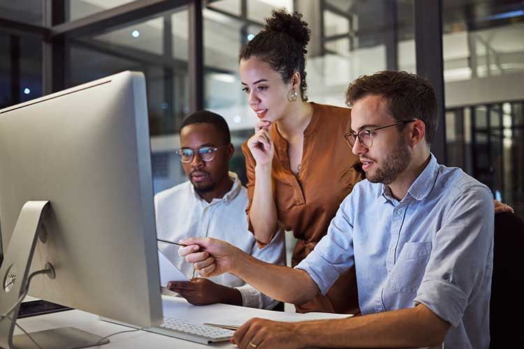 Three office workers looking at computer, man pointing, nighttime setting.