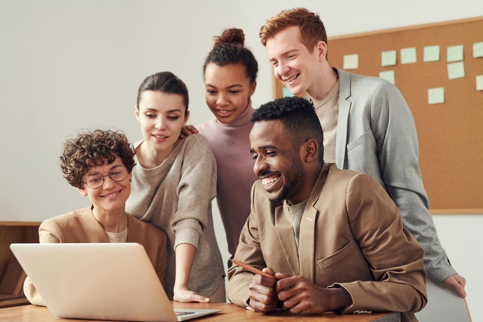 Five diverse colleagues around a laptop, smiling and focused in an office setting.