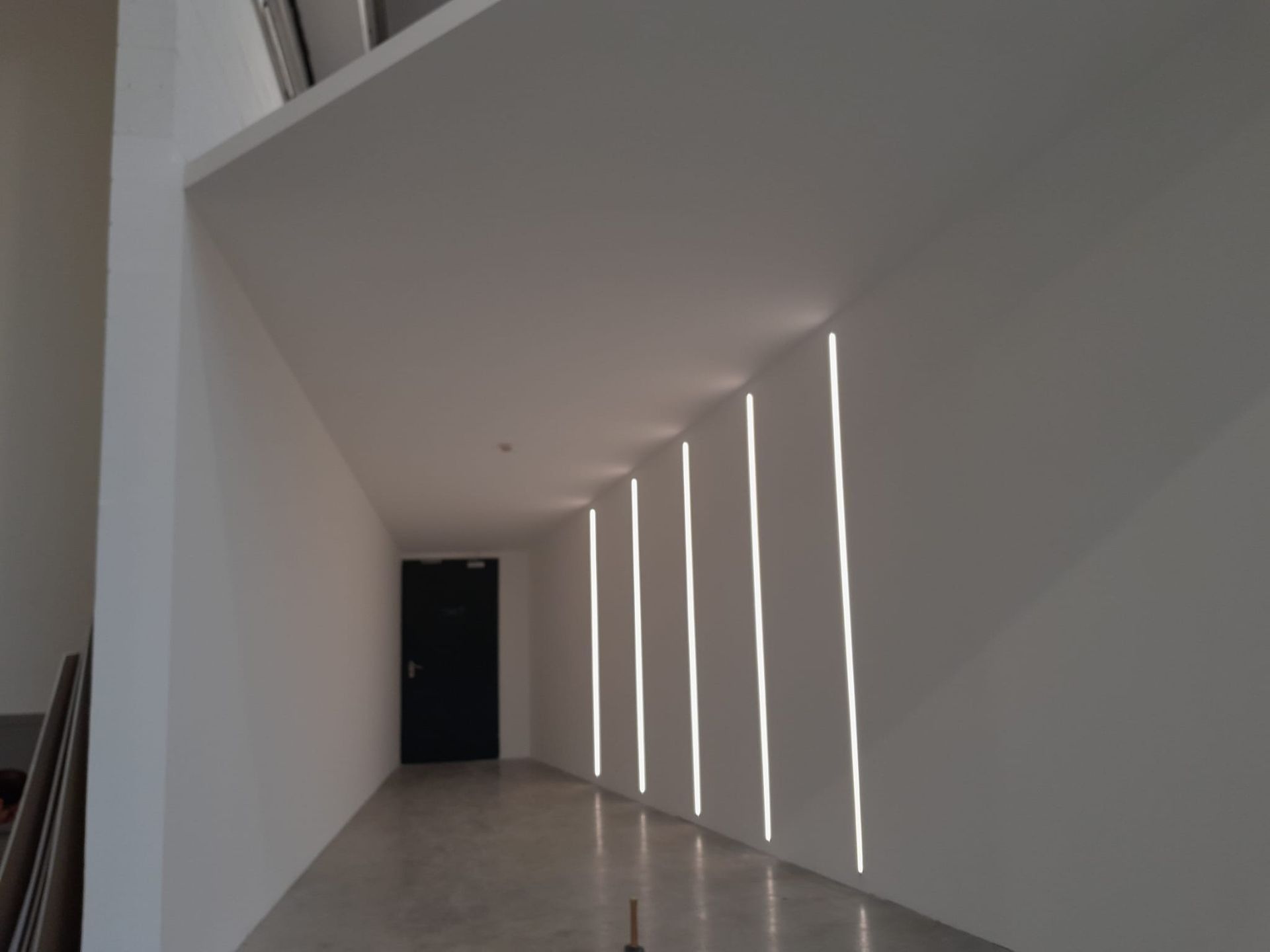 Corridoio bianco con luci lineari che illuminano la parete destra. In fondo c'è una porta scura.