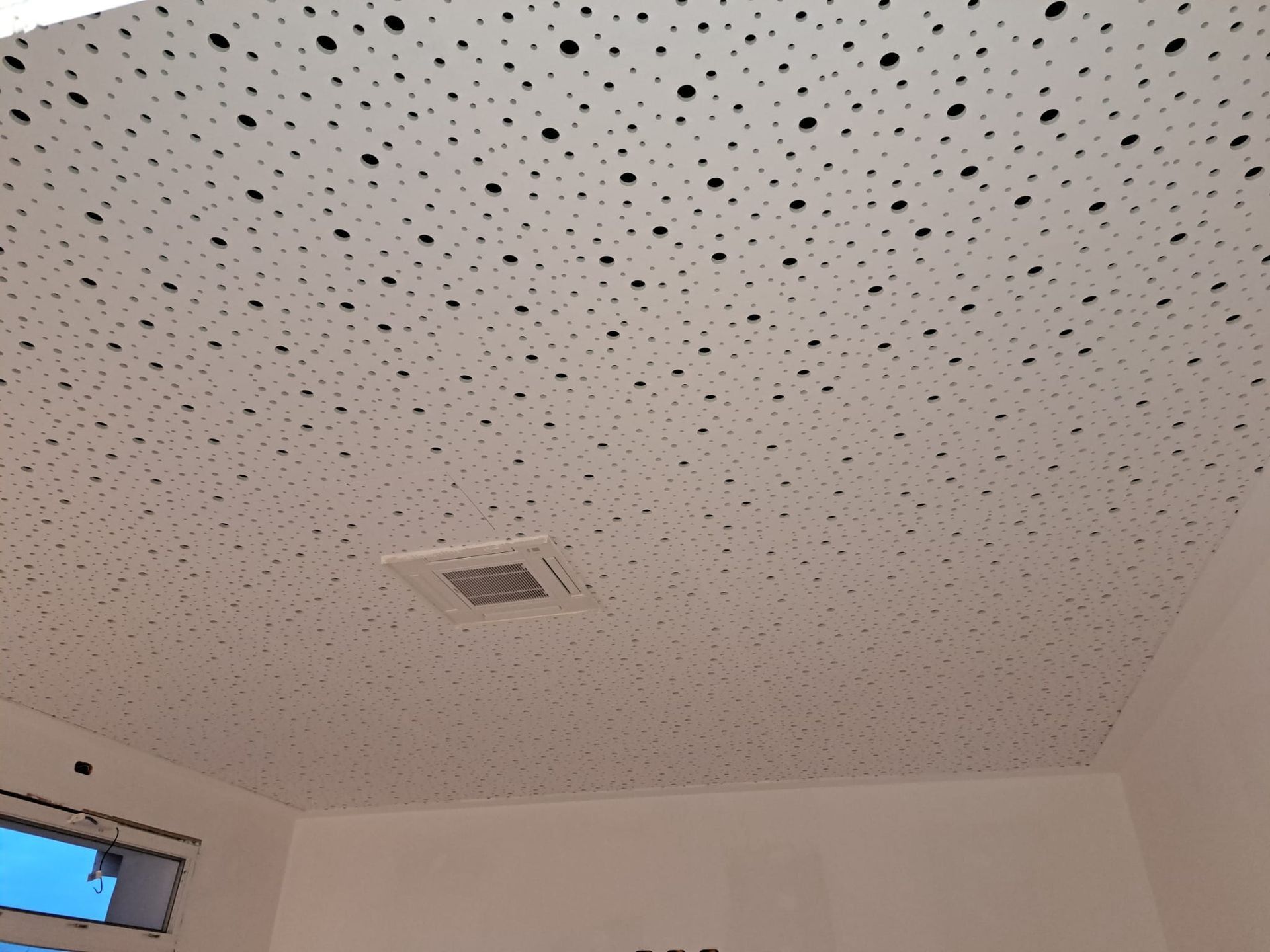 Soffitto bianco con piccoli fori neri, una presa d'aria quadrata e semplici pareti bianche.