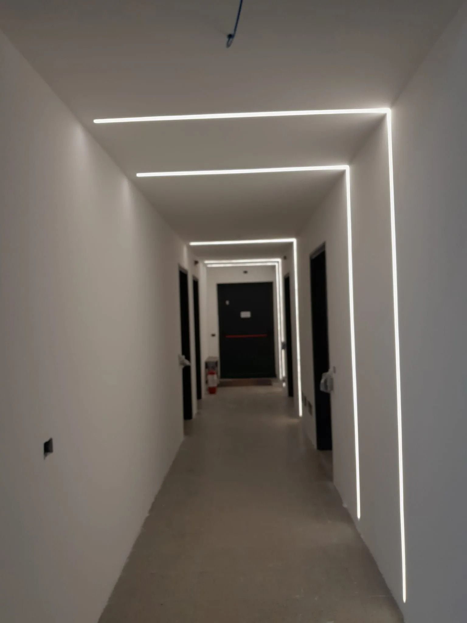 Corridoio con illuminazione a LED da incasso, pareti bianche, porte scure e un estintore.