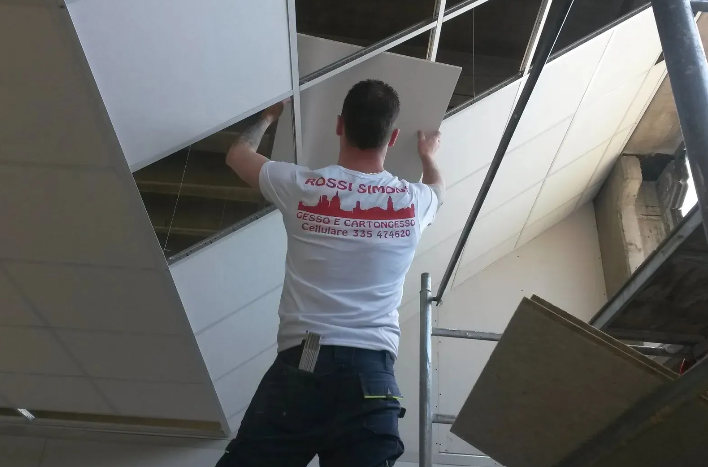 Uomo che installa i pannelli del soffitto di un edificio, utilizzando un'impalcatura. Camicia bianca e jeans.