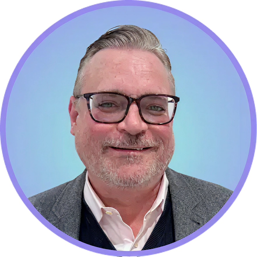 Man with glasses, graying hair, and beard, smiling, wearing a blazer over a sweater, set against a blue background.