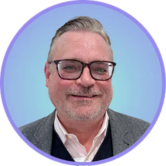 Man with glasses, graying hair, and beard, smiling, wearing a blazer over a sweater, set against a blue background.