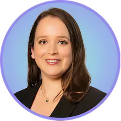 Woman with brown hair smiling, wearing a black blazer and necklace, against a blue background.