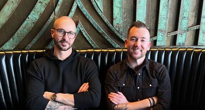 Two men sit side-by-side, arms crossed. One wears glasses and a black sweater; the other a black shirt and bracelets.