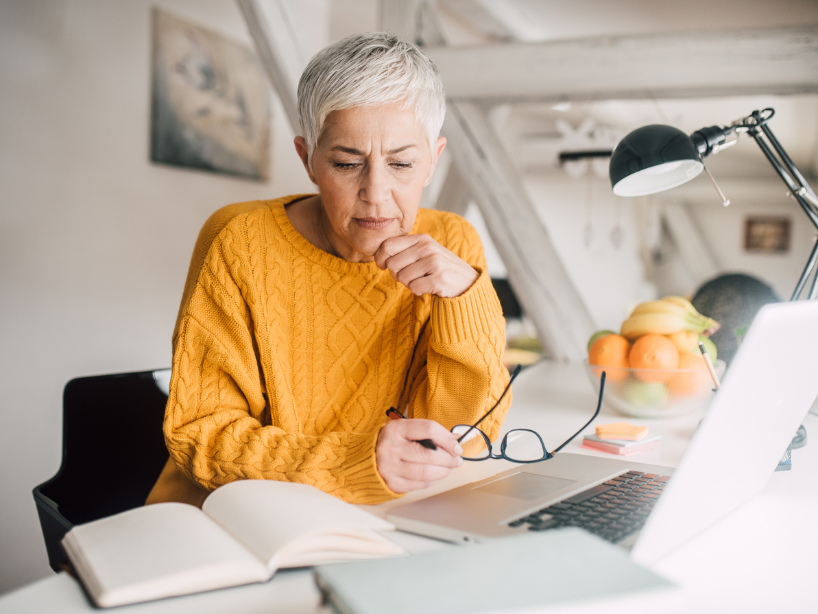 Woman with gray hair in a yellow sweater looks at laptop, holding glasses, notebook open, at a desk.