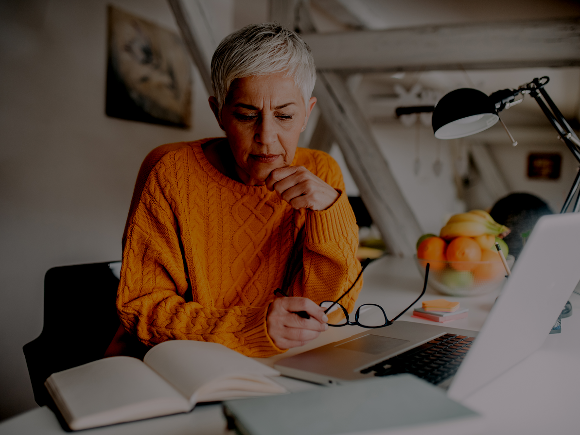 Woman with gray hair in an orange sweater, working at a desk with a laptop, glasses, and a notebook.