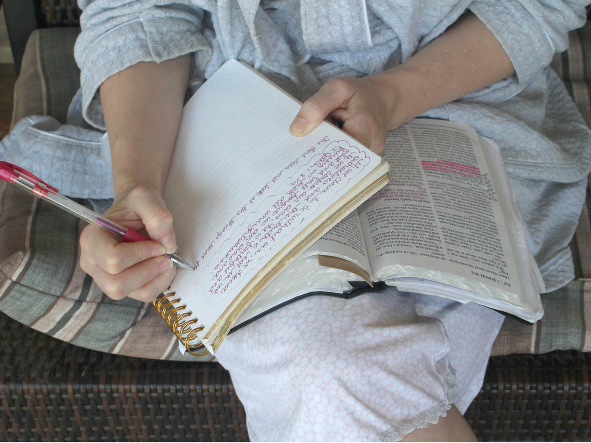 Woman sitting, writing in a notebook with a pen, Bible open on her lap. Light colors, close-up shot.