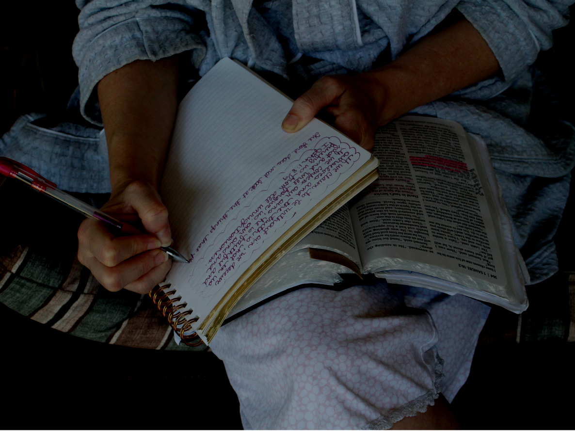 Person writing in a notebook, with an open book, on their lap.