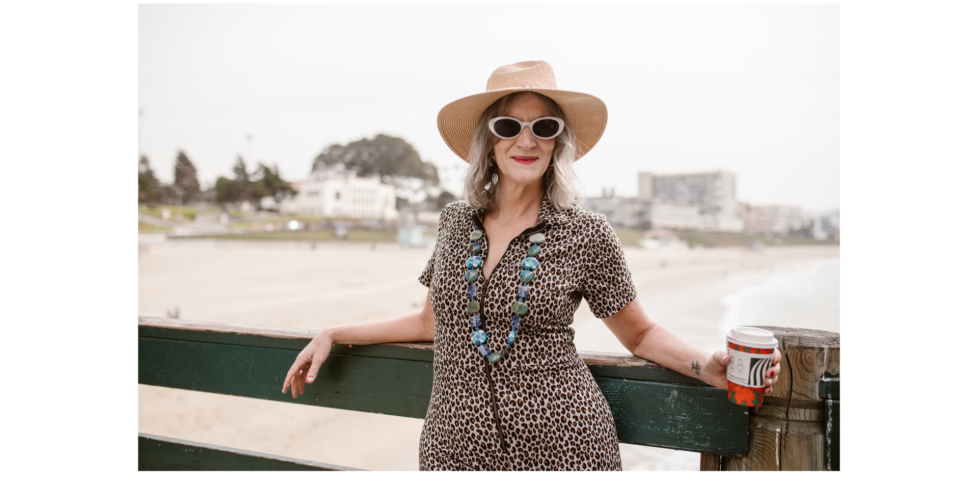 Woman in hat and sunglasses on pier, holding a cup, beach in the background.