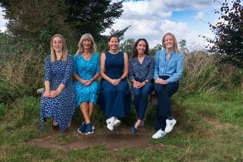 Five women sitting on a bench outdoors, smiling. Blue and patterned clothing, natural setting.