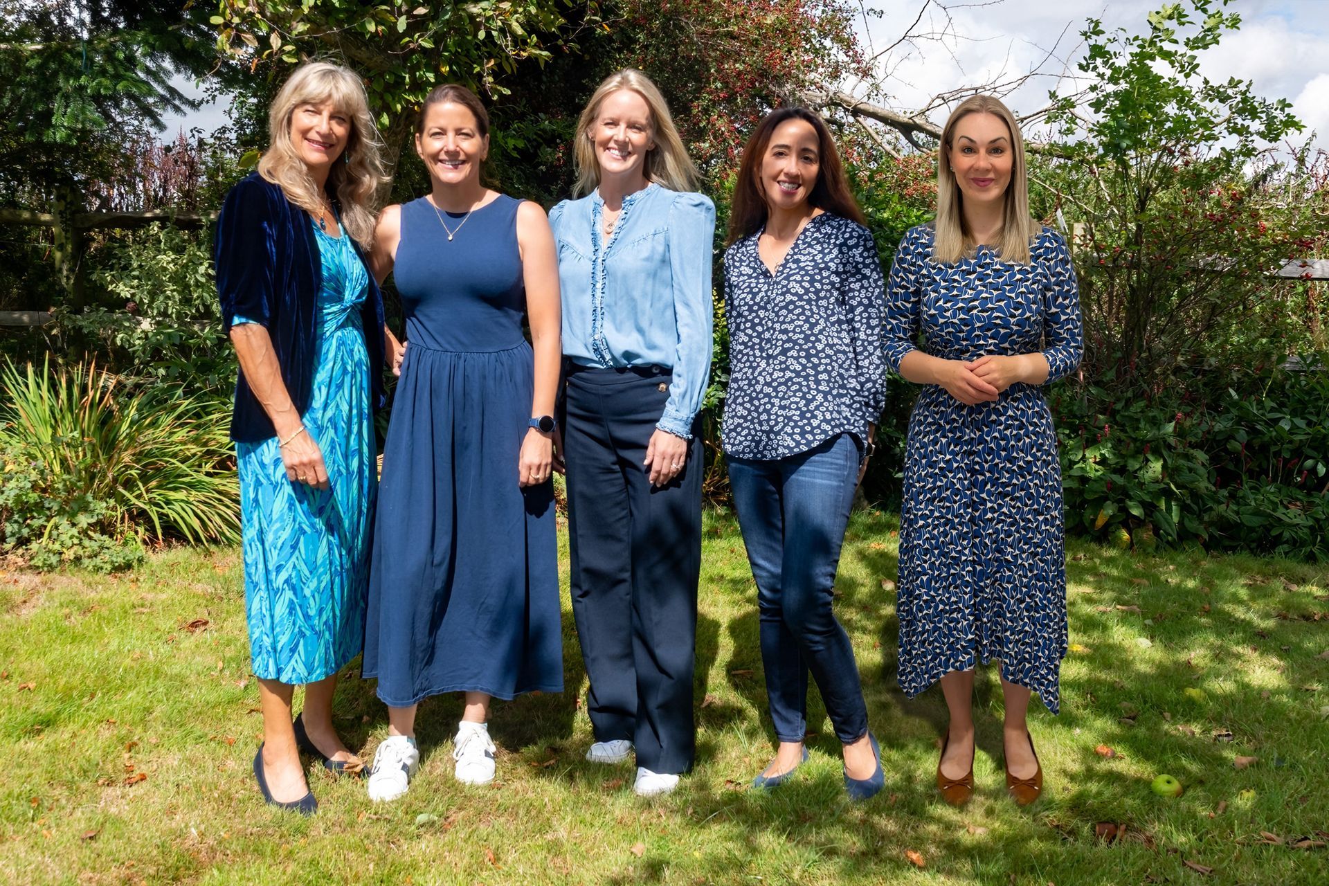 Five women in blue outfits pose outdoors on a sunny day, smiling.