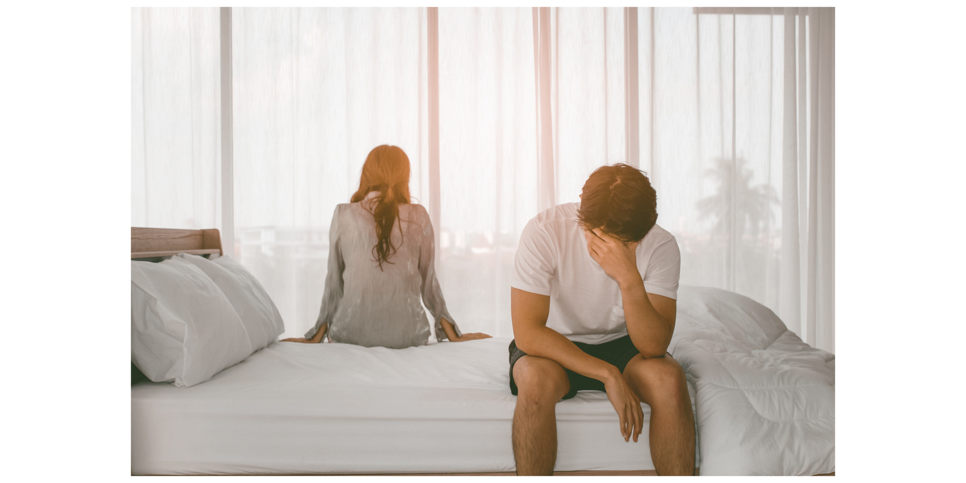 A couple sits on a bed, separated. The man is distraught, covering his face. Sunlight streams through a window.