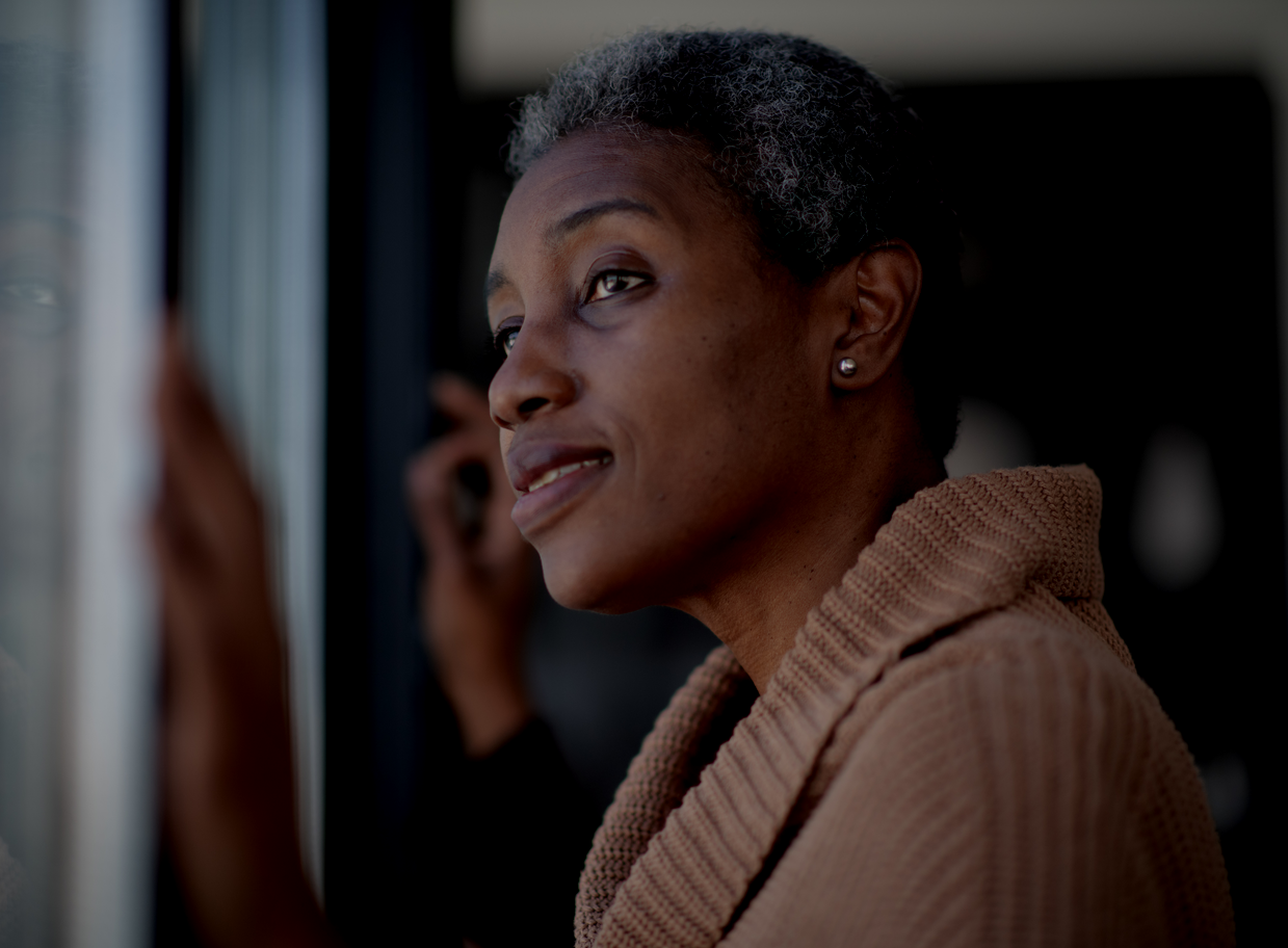 Woman with gray hair gazes out a window, wearing a tan sweater in a dimly lit room.