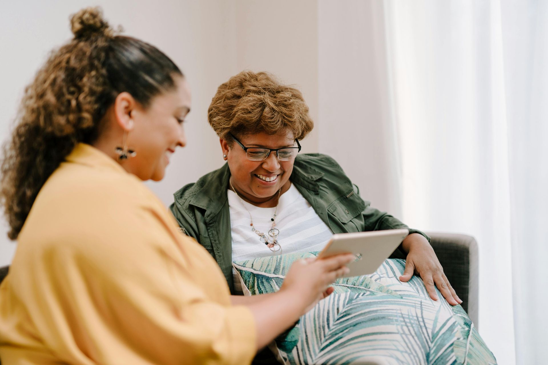 Woman in yellow shirt shows tablet to smiling woman wearing glasses. Both are seated indoors.